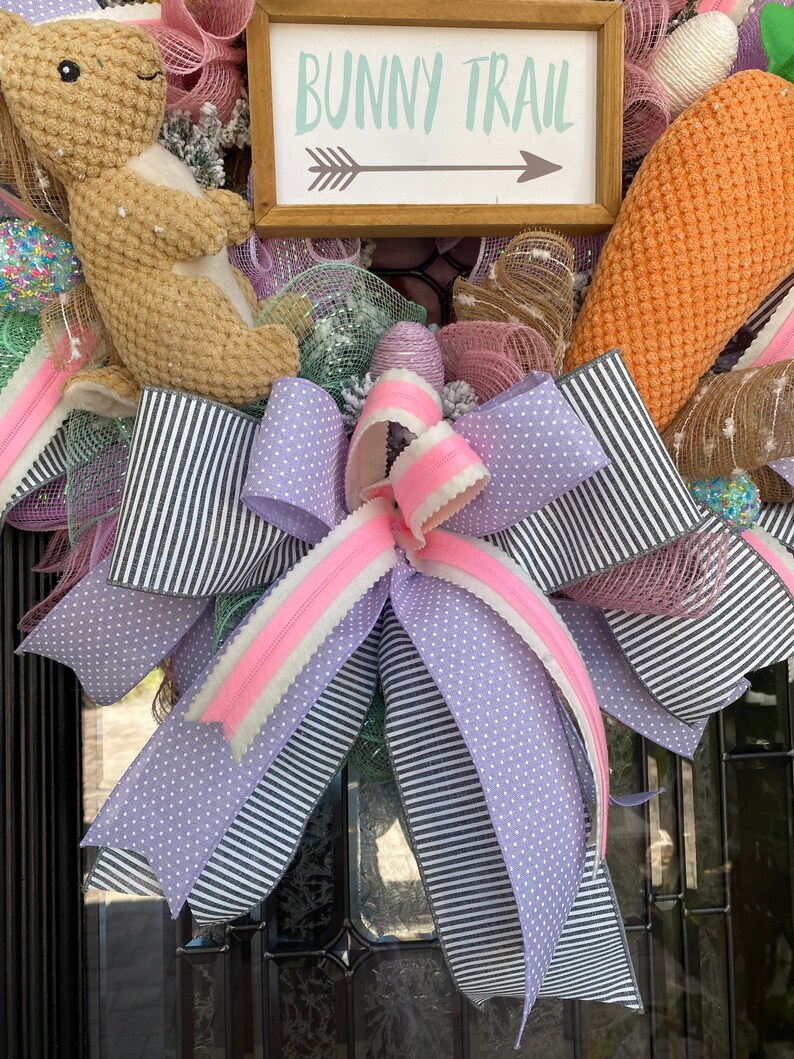 May include: Easter wreath with a brown bunny, carrot, and decorative ribbons. The wreath features a sign that reads "Bunny Trail" with an arrow. Ribbons are black and white striped, lavender polka dot, and pink.