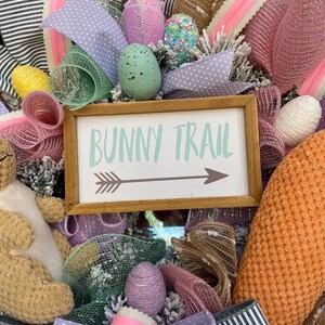 May include: A decorative Easter wreath with a wooden sign that reads "Bunny Trail" with an arrow. The wreath features pastel-colored ribbons, mesh, and decorative eggs in shades of pink, purple, and green. A plush bunny is also visible.