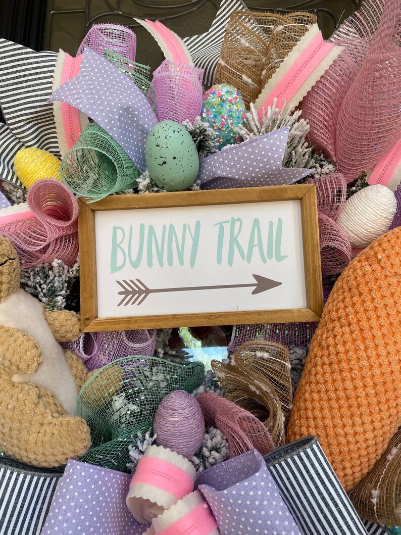 May include: A decorative Easter wreath with a wooden sign that reads "Bunny Trail" with an arrow. The wreath features pastel-colored ribbons, mesh, and decorative eggs in shades of pink, purple, and green. A plush bunny is also visible.