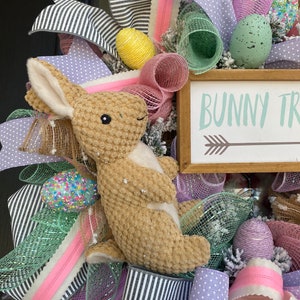 May include: Easter wreath with a plush bunny, decorative eggs, and ribbons in pastel colors. The wreath includes a wooden sign that reads "Bunny Trail" with an arrow. The design incorporates black and white striped ribbon.