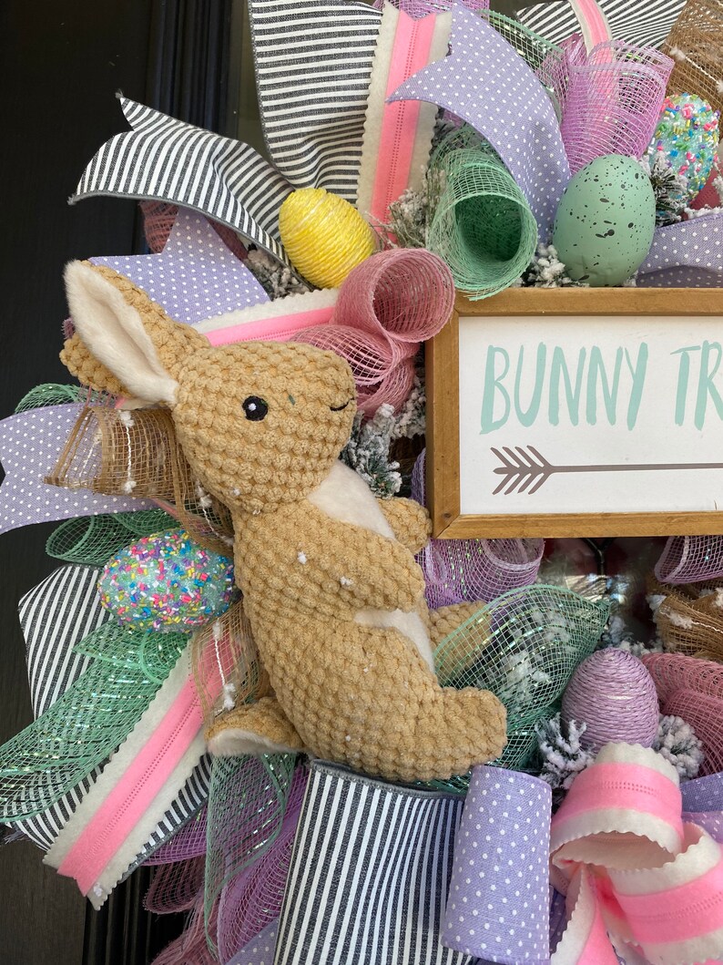 May include: Easter wreath with a plush bunny, decorative eggs, and ribbons in pastel colors. The wreath includes a wooden sign that reads "Bunny Trail" with an arrow. The design incorporates black and white striped ribbon.