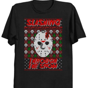 May include: Black t-shirt with a festive design featuring a white hockey mask with a red slash, surrounded by skulls and Christmas trees. The text reads "SLASING THROUGH THE SNOW" in red.