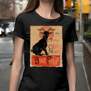 May include: Black t-shirt featuring a vintage-style graphic of a black goat with curved horns, set against a tan background with red accents. The text "TOURN&Eacute;E DE LA CH&Egrave;VRE NOIRE" and "Black Phillip" are also displayed.