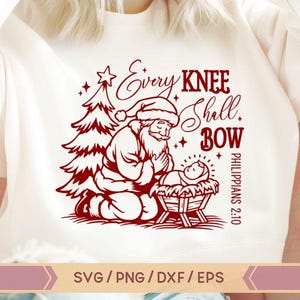 May include: Red and white Christmas-themed design featuring a kneeling Santa Claus with a baby Jesus in a manger. The text reads "Every knee shall bow Philippians 2:10".
