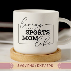 Op de afbeelding: Een witte keramische mok met een zwart-wit ontwerp met de tekst "Living that Sports Mom Life". De mok staat op een houten onderzetter.