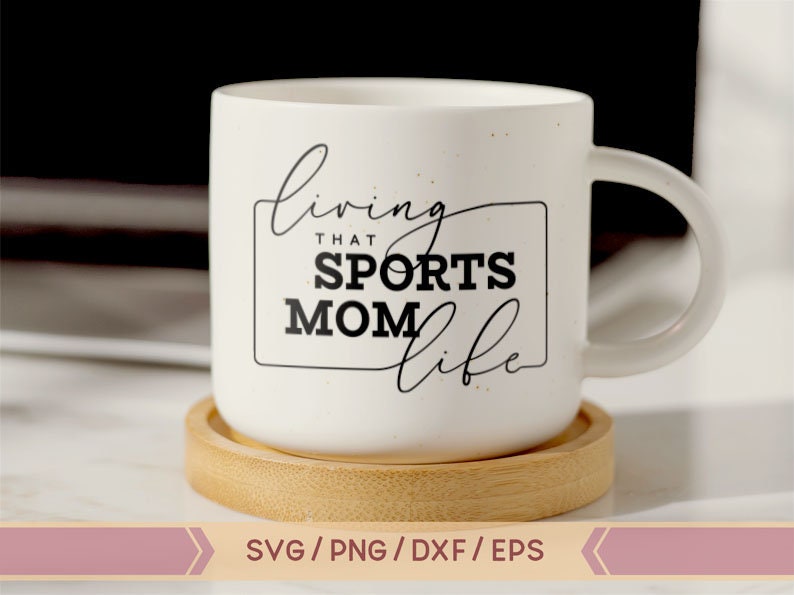 Op de afbeelding: Een witte keramische mok met een zwart-wit ontwerp met de tekst "Living that Sports Mom Life". De mok staat op een houten onderzetter.