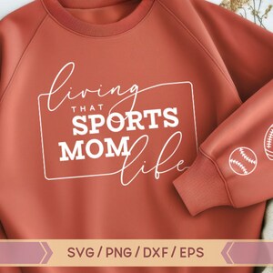Op de afbeelding: Een roestkleurig sweatshirt met de tekst "Living that Sports Mom Life" in wit. Het sweatshirt heeft een witte omtrek van een rechthoek rond de tekst. De mouw van het sweatshirt heeft witte contouren van een honkbal, voetbal en basketbal.