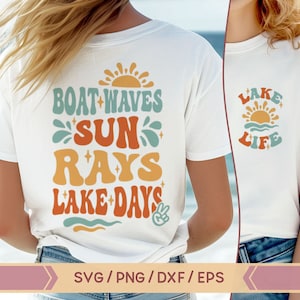Op de afbeelding: Twee witte T-shirts met een zomers ontwerp. Eén shirt zegt "Boat Waves Sun Rays Lake Days" met een vredesteken. Het andere shirt zegt "Lake Life" met een zon en golven.