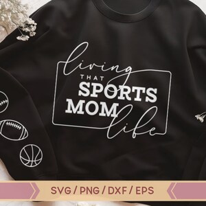Op de afbeelding: Zwart sweatshirt met witte tekst die "Living that Sports Mom Life" luidt. Het sweatshirt heeft een witte baseball, voetbal en basketbal afbeelding op de mouw.