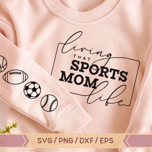 Op de afbeelding: Roze sweatshirt met de tekst "Living that Sports Mom Life" in zwarte cursieve letters. De mouw heeft een rij zwarte sportpictogrammen: een basketbal, een voetbal, een voetbal en een honkbal.
