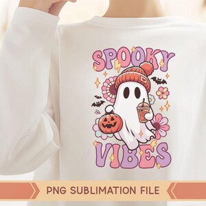May include: A white long-sleeved shirt with a pink, purple, and white graphic design featuring a ghost holding a pumpkin and a cup of coffee. The text "Spooky Vibes" is written in pink and purple letters.