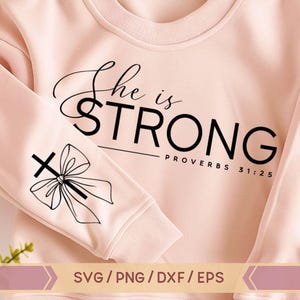 May include: A pink sweatshirt with the text "She is STRONG - Proverbs 31:25" printed on the front. The left sleeve has a black and white illustration of a cross with a bow.