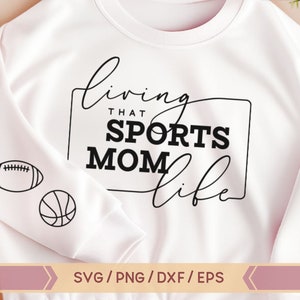Op de afbeelding: Wit sweatshirt met zwarte tekst die "Living that Sports Mom Life" luidt, met een zwarte omtrek rond de tekst. Aan de linkerkant van het sweatshirt staan drie zwarte en witte sportpictogrammen: een honkbal, een voetbal en een basketbal.