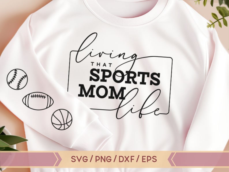 Op de afbeelding: Wit sweatshirt met zwarte tekst die "Living that Sports Mom Life" luidt, met een zwarte omtrek rond de tekst. Aan de linkerkant van het sweatshirt staan drie zwarte en witte sportpictogrammen: een honkbal, een voetbal en een basketbal.