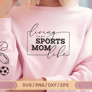 Op de afbeelding: Roze sweatshirt met de tekst "living that sports mom life" in zwarte letters. De mouw heeft drie zwart-witte iconen van een honkbal, voetbal en voetbal.