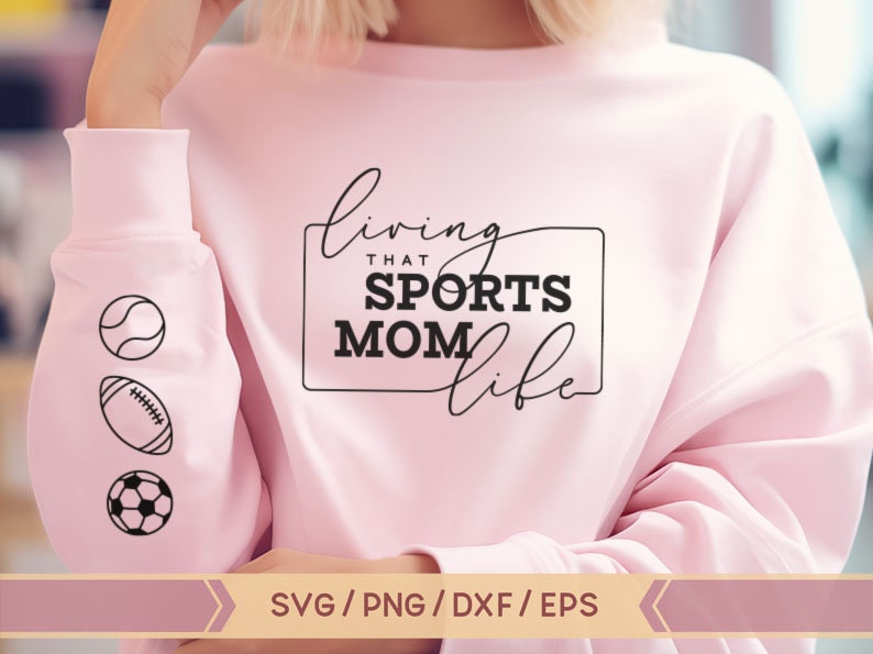 Op de afbeelding: Roze sweatshirt met de tekst "living that sports mom life" in zwarte letters. De mouw heeft drie zwart-witte iconen van een honkbal, voetbal en voetbal.