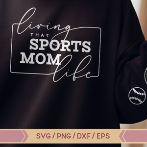 Op de afbeelding: Zwart sweatshirt met witte tekst die "Living that Sports Mom Life" luidt. Het sweatshirt heeft witte contouren van sportballen op de mouw.