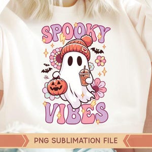 May include: A white t-shirt with a pink and purple graphic design featuring a cartoon ghost wearing a hat and holding a pumpkin and a cup. The text "Spooky Vibes" is written in pink and purple letters.