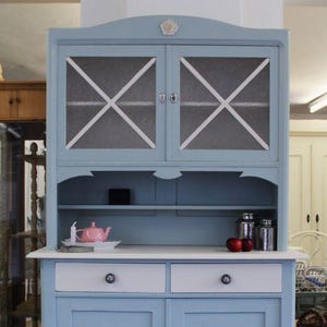 Könnte beinhalten: Ein hellblaues Vintage-Schrank mit Glastüren und weißen Akzenten. Der Schrank hat zwei Schubladen und zwei untere Türen. Eine rosa Teekanne und zwei rote Äpfel stehen auf der Arbeitsplatte. Ein charmantes Möbelstück.