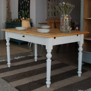 Könnte beinhalten: Rechteckiger Holztisch mit einer Tischplatte aus Naturholz und einem weiß lackierten Untergestell. Der Tisch hat vier gedrechselte Beine und eine Schublade. Eine Vase mit Trockenblumen steht auf dem Tisch, zusammen mit drei weißen Schalen.