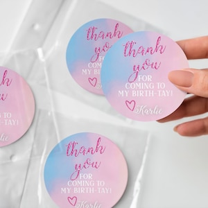 Peut inclure: Trois autocollants ronds roses et bleus de remerciement avec le texte "thank you for coming to my birth-tay!" et le nom "Karlie" en police de caract&egrave;res rose paillet&eacute;e.