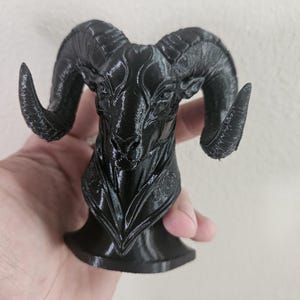 May include: A black ram sculpture with detailed horns and facial features. The ram is mounted on a small, round base. The sculpture is held in a hand against a plain background.