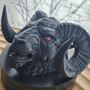 Puede incluir: Escultura de calavera de carnero negro con texturas detalladas y cuernos curvados. La escultura tiene una expresión amenazante con dientes visibles y un solo ojo rojo. Montada sobre una base circular negra, ideal para exhibir.