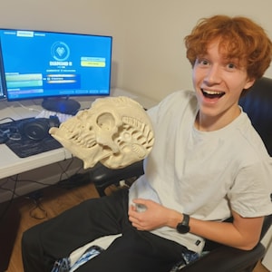 May include: A beige, intricately detailed skull sculpture. The skull has a textured surface with visible bone structure and openings. The sculpture is held by a person in a white t-shirt and black pants.