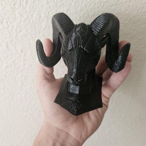 May include: A black ram sculpture with detailed horns and a textured surface. The ram's head is mounted on a geometric base. The sculpture is held in a hand against a neutral background. The sculpture is likely a decorative item.