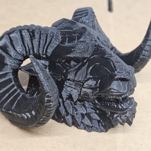 Könnte beinhalten: Eine schwarze, 3D-gedruckte Widderkopf-Skulptur. Die Skulptur weist detaillierte Texturen und markante, gebogene Hörner auf. Der Widderkopf ist ein dunkles, mattes Schwarz mit sichtbaren Schichtlinien. Es ist ein dekorativer Artikel.