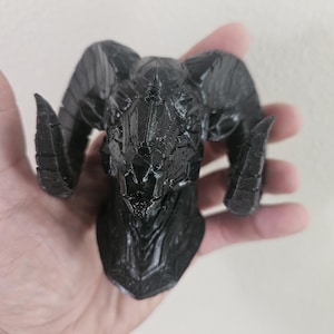 Könnte beinhalten: Ein schwarzer, 3D-gedruckter Widder-Schädel mit großen, gebogenen Hörnern. Der Schädel hat eine detaillierte, strukturierte Oberfläche und wird in der Hand einer Person gehalten. Der Widder-Schädel ist ein dekorativer Gegenstand.