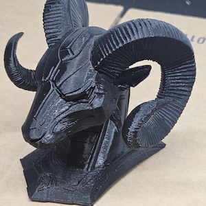 May include: A black 3D-printed sculpture of a ram's head. The sculpture has detailed horns, a textured surface, and a geometric design. The ram's head is mounted on a rectangular base, showcasing a modern, artistic aesthetic.