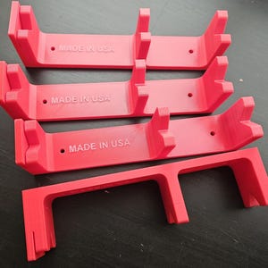 Op de afbeelding: Verschillende helderrode, 3D-geprinte beugels met een uniek ontwerp. Elke beugel heeft een rechthoekige basis met twee verticale steunen en een gebogen bovenkant. De tekst "MADE IN USA" is op elke beugel gedrukt.