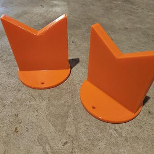 Puede incluir: Dos sujetalibros de color naranja brillante en forma de V. Cada sujetalibros tiene una base semicircular con un pequeño agujero. Los sujetalibros están hechos de un material liso y están diseñados para estar de pie.