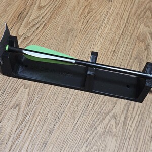 Op de afbeelding: Een zwarte pijlhouder met één pijl. De pijl heeft een groene en witte pijlschacht. De houder is rechthoekig met een verhoogde rand en steunen. De tekst "MADE IN..." is zichtbaar op de houder.