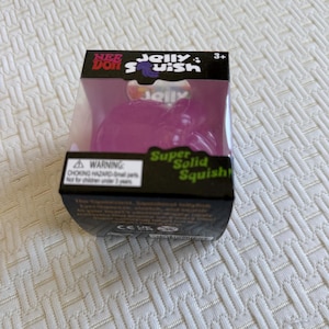 May include: A NeeDoh Jelly Squish toy in a black cardboard box with a clear plastic window. The toy is a translucent purple, squishy, jellyfish-shaped object. The packaging features the text NeeDoh Jelly Squish, Super Solid Squish, and a warning.