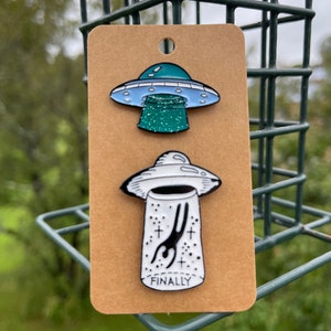 May include: Two enamel pins on a brown card. The top pin is a blue and white UFO with a green beam. The bottom pin is a white and black UFO with a person being beamed up, and the word "FINALLY".