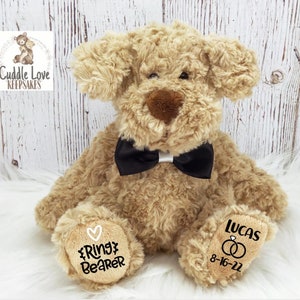 May include: A beige plush dog wearing a black bow tie. The dog has a heart and the words "Ring Bearer" embroidered on one paw and the name "Lucas" and the date "8-16-22" with two rings embroidered on the other paw.