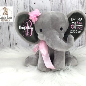 May include: Gray plush elephant stuffed animal with a pink bow and a pink flower on its head. The elephant has a black felt patch on its ear with the name "Berkeley" and birth stats: "12-11-18 6 pounds 8 oz 19 3/4 in 12:03 am"