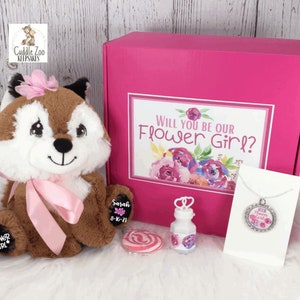 May include: Pink gift box with a flower girl theme. The box contains a plush fox toy, a necklace, a bottle of bubbles, a lollipop, and a personalised tag. The box has the text "Will you be our Flower Girl?" on the front.