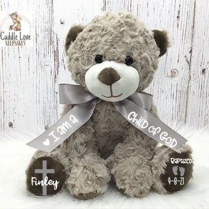May include: A gray plush teddy bear with a gray ribbon tied around its neck. The ribbon has the text "I am a Child of God" printed on it. The bear's paws have a cross and the name "Finley" printed on one paw and the text "Baptized 9-8-21" printed on the other paw.