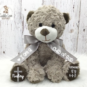 May include: A gray teddy bear with a white face and brown nose. The bear is wearing a gray ribbon with the text "I am a Child of God" and a cross on one paw. The other paw has the text "Finley 9-8-21".