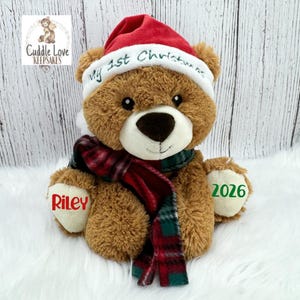 May include: A brown plush teddy bear wearing a red Santa hat with the embroidered text My 1st Christmas and a red and green plaid scarf. The bear has cream-colored paw pads with the name Riley in red and the year 2026 in green embroidered on them.