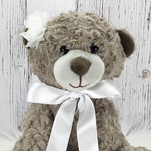 May include: A plush brown teddy bear with a white satin bow and a white flower on its ear.