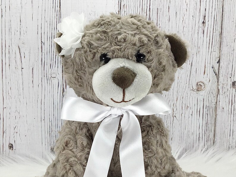May include: A plush brown teddy bear with a white satin bow and a white flower on its ear.