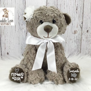 May include: A grey plush teddy bear with a white satin bow and a white flower on its head. The bear has embroidered text on its paws that reads "Flower Girl" and "Sarah 8-16-22".