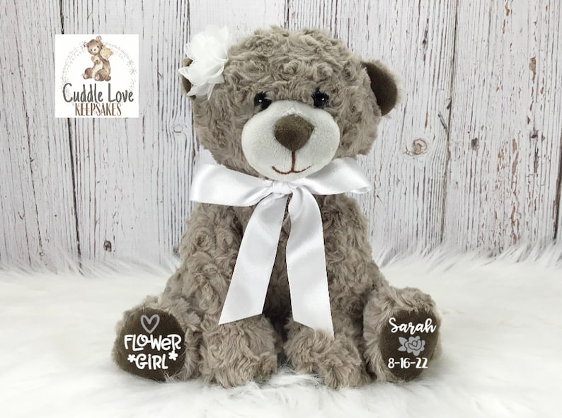May include: A grey plush teddy bear with a white satin bow and a white flower on its head. The bear has embroidered text on its paws that reads "Flower Girl" and "Sarah 8-16-22".