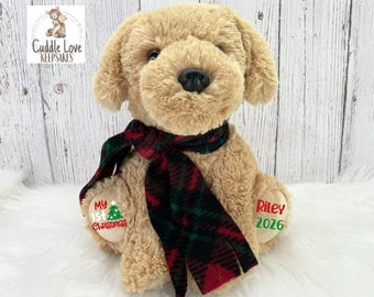 May include: A tan plush dog toy wearing a red and green plaid scarf. The left foot pad features the text "My 1st Christmas" with a small tree graphic, and the right foot pad displays the name "Riley" above the year "2026" in red and green.