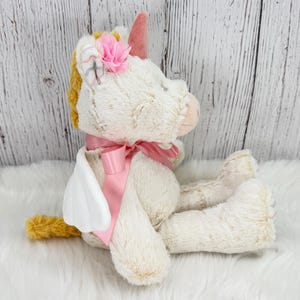 Pode incluir: Um brinquedo de pel&uacute;cia de unic&oacute;rnio creme e dourado com uma fita, chifre e flor rosa. O unic&oacute;rnio tem asas brancas e est&aacute; sentado em uma superf&iacute;cie branca e fofa. O fundo &eacute; um painel de madeira branco.