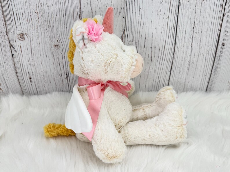 Pode incluir: Um brinquedo de pel&uacute;cia de unic&oacute;rnio creme e dourado com uma fita, chifre e flor rosa. O unic&oacute;rnio tem asas brancas e est&aacute; sentado em uma superf&iacute;cie branca e fofa. O fundo &eacute; um painel de madeira branco.